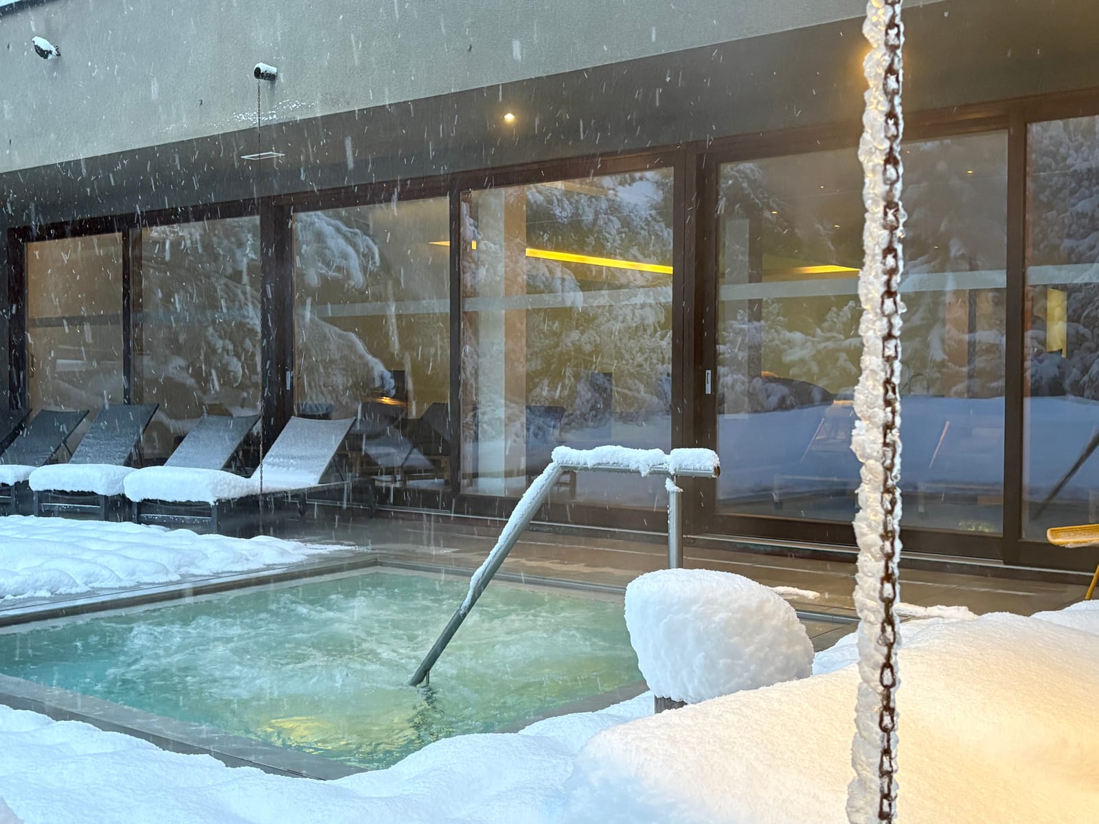 Außenpool im Schnee mit dampfendem Wasser hinter Glasfront, Liegen innen – MONTAFONLodge