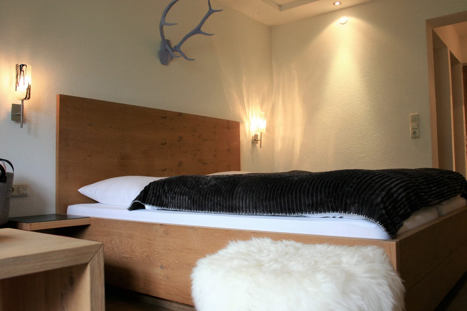 Doppelbett mit dunkler Tagesdecke und Schaffell, Deko-Geweih an der Wand – MONTAFONLodge