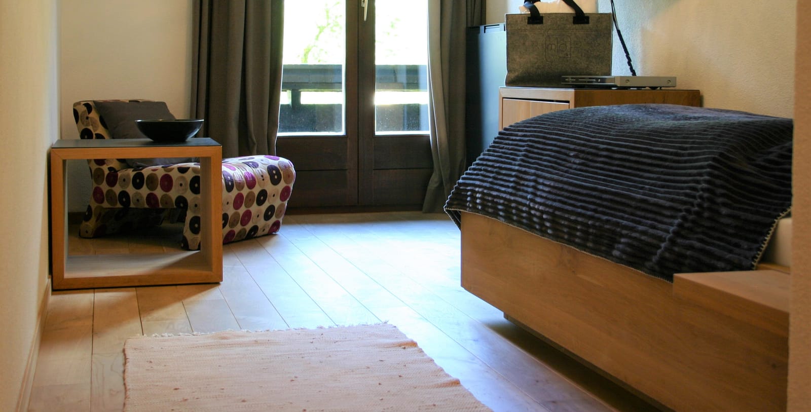 Einzelbett mit blauer Kordsamt-Tagesdecke, gepolsterter Hocker, bodentiefe Balkontür – MONTAFONLodge
